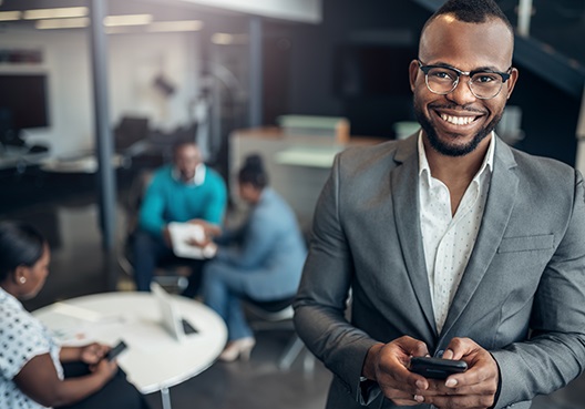 A guy smiling at the camera while holding his cellphone with a group of people sitting in the background