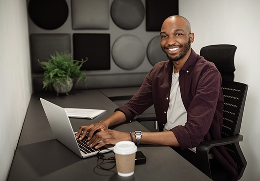 A man sitting in his home office with his fingers on his laptop, and a takeaway cup of coffee, smiling at the camera