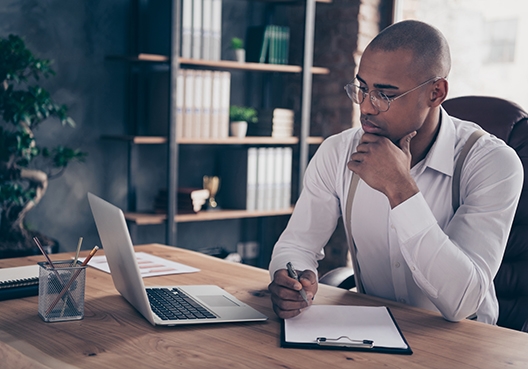man sitting behind his desk looking at his laptop holding a pen