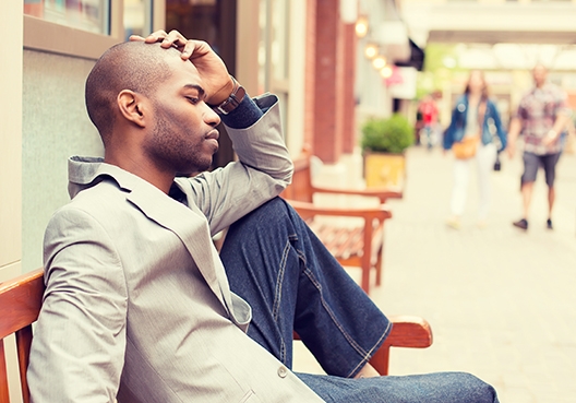 man sitting on a bench with his one leg bent with one foot on the bench and his arm leaning on his knee covering his face