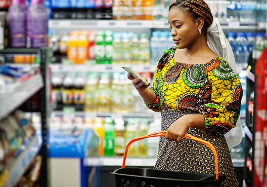 A young woman standing in an aisle in a supermarket holding a shopping basket while looking at her cellphone